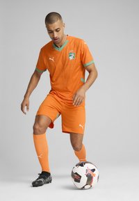Equipación de fútbol naranja con patrón texturizado, acentos verdes y logo. Jugador con botas negras controla un balón de fútbol negro y blanco.