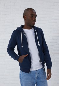 Felpa blu navy con zip, drawstrings bianchi, tasche frontali e polsini a coste, indossata sopra una T-shirt bianca e abbinata a jeans di un blu chiaro.