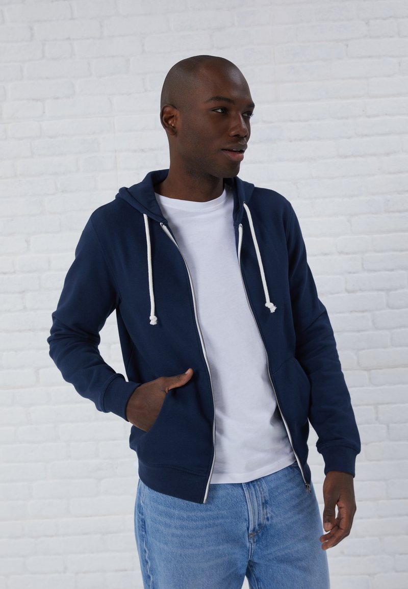 Felpa blu navy con zip, drawstrings bianchi, tasche frontali e polsini a coste, indossata sopra una T-shirt bianca e abbinata a jeans di un blu chiaro.