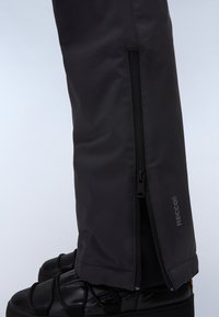 Zwarte waterdichte gaiters met zijritsen, gemaakt van synthetisch materiaal, met RECCO-branding, gedragen bovenop zwarte geïsoleerde laarzen.