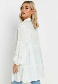 Blusa bianca leggera con design fluido, maniche lunghe e ampie, e dettagli in pizzo. Disponibile con una vestibilità ampia e sezioni a strati.