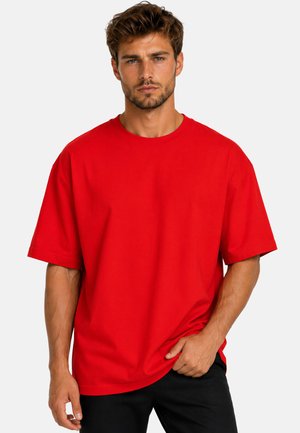 Uomo con capelli castani corti e barba che indossa una maglietta rossa oversize tinta unita e pantaloni neri, in piedi davanti a uno sfondo chiaro.