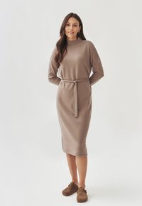 Robe en tricot à manches longues en tissu marron doux, avec un col roulé côtelé et une ceinture à la taille. Longueur midi avec fentes latérales, associée à des sabots marron.