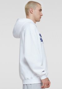 Witte hooded sweatshirt met ribgebreide manchetten en onderrand. De voorkant heeft blauwe geborduurde tekst. De stof lijkt zacht en lichtjes gestructureerd.