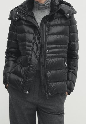 Person trägt eine schwarze gesteppte Daunenjacke über einem grauen Rollkragenpullover und dunkelgrauen Hosen, Hände in den Taschen, die Jacke teilweise geöffnet.