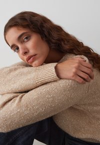 Pull en maille beige clair avec des speckles texturés, doté de poignets côtelés et d'une coupe décontractée, porté dans une pose décontractée sur un fond uni.