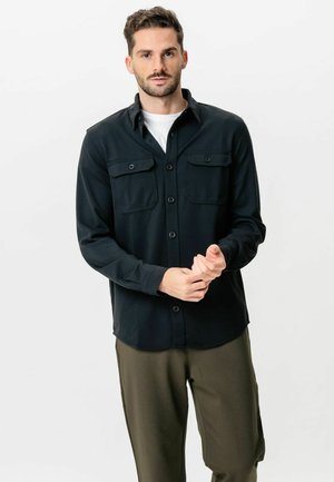 Mann trägt ein schwarzes, geknöpftes Hemd über einem weißen Unterhemd, justiert seinen rechten Ärmel und steht vor einem schlichten, hellen Hintergrund.