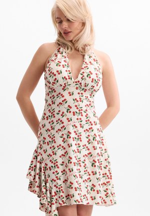 Mujer con cabello rubio que lleva un vestido blanco sin mangas con estampado rojo de cerezas y un escote halter, de pie frente a un fondo blanco.