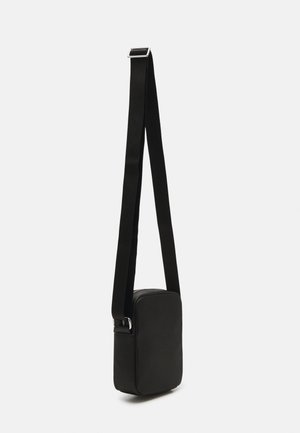 Sac bandoulière rectangulaire noir texturé avec sangle ajustable et boucle argentée sur un fond clair uni.