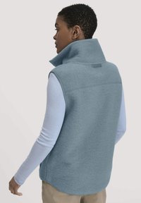 Helles blaues Fleeceweste mit hohem Kragen, seitlichen Taschen und abgerundetem Saum, getragen über einem hellen Langarmoberteil. Glatte Textur, lässiges Design.