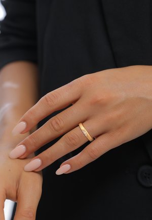 Gold-tone ring with three small clear stones, worn on a hand with light pink, oval-shaped nails. Smooth band design against a dark background.