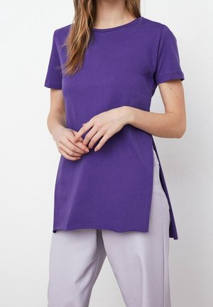 Frau trägt ein kurzärmeliges lila Tunika-Oberteil mit Seitenschlitzen, kombiniert mit hellen Hosen vor einem schlichten Hintergrund.