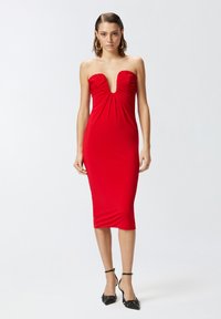 Crvena haljina bez naramenica s dubokim V-izrezom, naboranim korzetom i uskim siluetom. Glatka tekstura, haljina do koljena, naglašena crnim potpeticama.