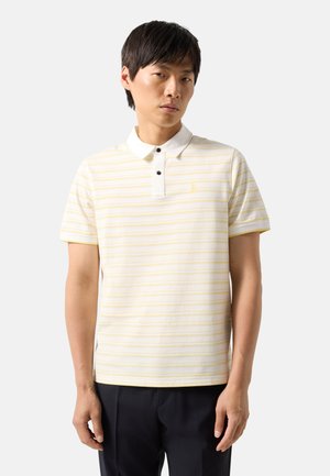 Homme portant un polo blanc à manches courtes avec de fines rayures horizontales jaunes et un pantalon noir, debout devant un fond clair uni.