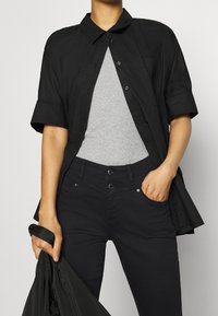 Persona con una camisa negra de manga corta y botones sobre una camiseta sin mangas gris y pantalones negros, sosteniendo una chaqueta negra con la mano izquierda y con la mano derecha en el bolsillo.