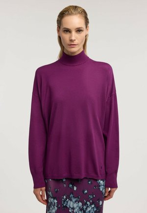 Femme aux cheveux blonds portant un pull à col roulé violet à manches longues et une jupe à motif floral sur un fond uni.