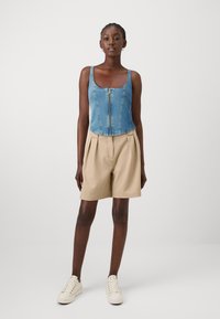 Top in denim con scollo a cuore e chiusura lampo frontale, abbinato a pantaloni corti beige a pieghe e sneakers color crema. Design semplice, texture lisce e colori a contrasto.
