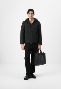 Sort puffer-jakke med lynlås foran og grøn foring, båret over en grå skjorte. Sorte bukser og en struktureret sort taske fuldender outfittet.