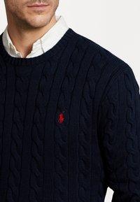 Marinblå kabelstickad tröja med ribbad rund halsringning och en liten röd logga på bröstet. Mjukt, texturerat tyg.