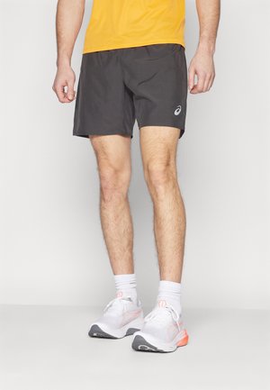 Homme portant un short de sport noir, des chaussettes blanches et des chaussures de course blanches avec des touches orange, debout devant un fond uni.