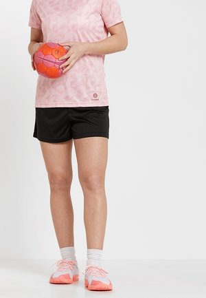 Osoba w różowym sportowym T-shircie, czarnych szortach, białych skarpetkach i różowo-białych sportowych butach trzymająca oburącz pomarańczowo-różową piłkę do piłki ręcznej.