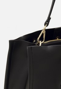 Bolso de mano de cuero negro con una textura suave. Presenta un zipper dorado, un accesorio de broche y un diseño de apertura superior.