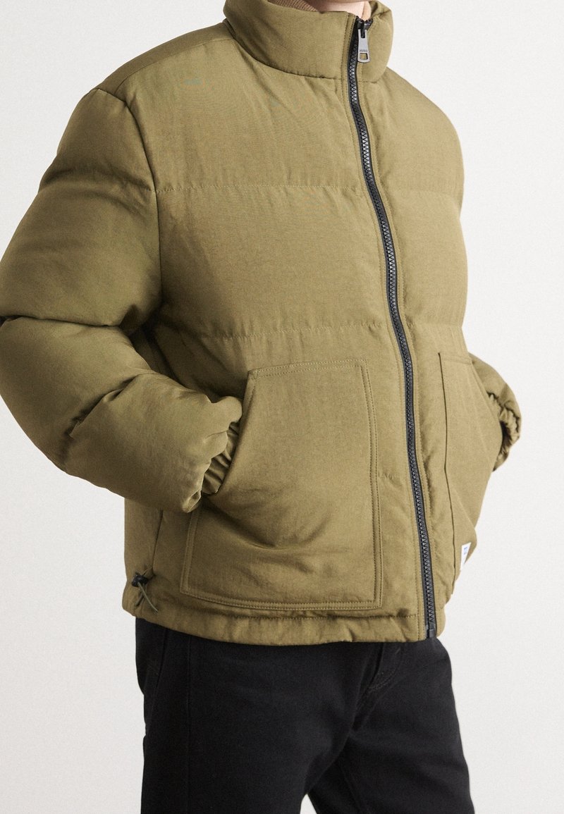 Homme portant une veste matelassée vert olive avec une fermeture éclair à l’avant et les mains dans les poches avant carrées, assortie à un pantalon noir.