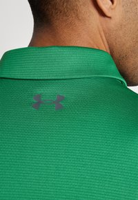 Zaļš polo krekls ar strukturētu audumu, ar melnu logo aizmugures kakla daļā un klasisku apkakles dizainu.