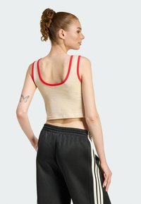 Top corto senza maniche in beige con bordatura rossa; abbinato a pantaloni della tuta neri con strisce bianche lungo i lati. Tessuto morbido, vestibilità comoda.