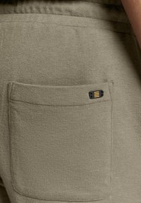 Olijfgroene joggers van textuurstof, met een achterzak en een klein zwart label met gouden accenten.