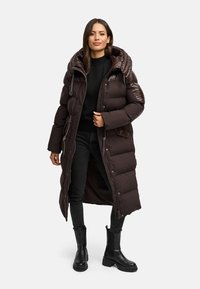 Lång mörkbrun puffjacka med quiltat mönster, överdimensionerad huva och glansiga detaljer. Bärs över en svart tröja och åtsittande jeans, ihop med svarta stövlar.