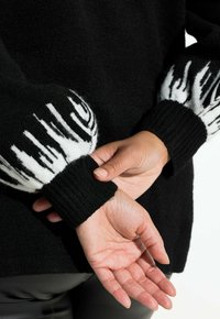 Jersey negro con puños acanalados, que presenta un patrón de llamas blancas en los extremos de las mangas. La tela parece suave con un acabado texturizado.