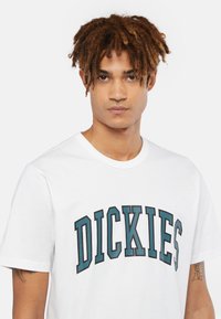 Vit bomullströja med en stor "DICKIES" logotyp i fet teal- och svart text. Korta ärmar och en klassisk rundad halsringning.