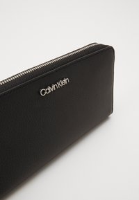 Juodas odinis piniginė su šiurkščiu paviršiumi, sidabrinė užtrauktukas ir iškilus "Calvin Klein" logotipas priekyje. Stačiakampio formos.