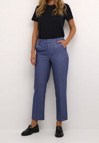 Donna vestita con una maglietta nera a maniche corte, pantaloni blu a vita alta con gamba dritta e mocassini neri, in piedi con una mano in tasca.