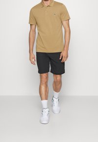 Béžové polo tričko s malým zeleným logem, černé šortky a bílé tenisky. Látka vypadá hladká a má ležérní design.