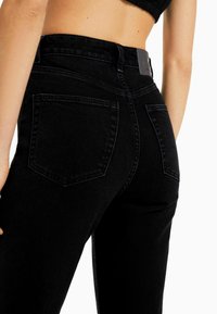 Pantalones vaqueros de mezclilla negra con cintura alta, que presentan una textura suave, diseño de cinco bolsillos y costuras a tono. La vista trasera muestra las formas de los bolsillos.