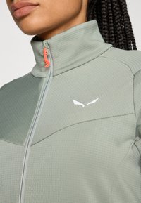 Lysegrønn zip-up jakke med høy krage, teksturert stoff, kontrastfarget oransje glider, og en hvit logo på brystet.