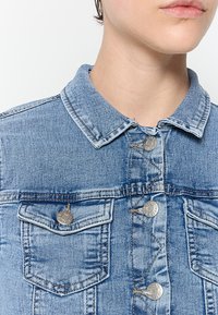 Ljusblå denimjacka med krage, silverfärgade tryckknappar och två bröstfickor. Har en klassisk, strukturerad tygdesign.