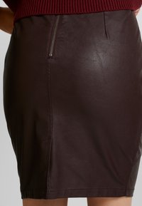 Gros plan sur une jupe crayon en cuir marron foncé avec un détail de fermeture éclair visible à la taille et de légères rides sur le tissu.