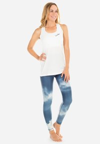 Weißes Tanktop mit runder Ausschnittsform und kleinem Logo, kombiniert mit blauen Leggings mit einem wolkenartigen Muster. Beides ist figurbetont und elastisch.