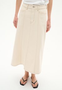 Beige Denimrock mit gerader Silhouette, der horizontale Nahtdetails und seitliche Taschen aufweist. Kombiniert mit schwarzen Flip-Flops.