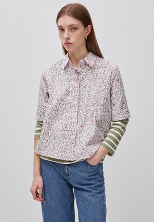 Junge Frau mit glattem braunem Haar trägt ein geblümtes Kurzarm-Hemd über einem langärmligen grün-weiß gestreiften Shirt und blaue Jeans.