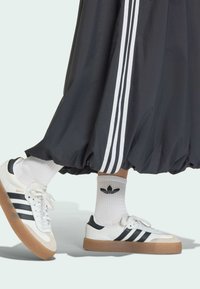 Topánky Adidas Samba v bielej a čiernej farbe s gumovou podrážkou, skombinované s bielymi členkovými ponožkami s čiernym logom, pod šedou splývavou sukňou.