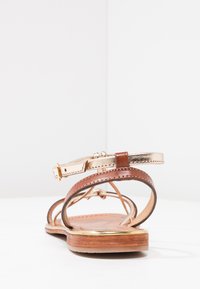 Sandalo in pelle marrone con una cinghia metallica dorata, suola piatta e chiusura con fibbia. Presenta un design minimalista e cuciture a vista.