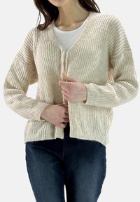 Chunky crèmekleurige cardigan in een textuurgebreide stof met een V-hals en een striksluiting aan de voorkant, gecombineerd met een wit T-shirt en donkere jeans.