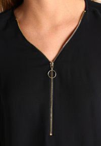 Black blouse featuring a gold zipper at the neck, with a circular pull. The fabric appears smooth and lightweight, with a v-neck design.
