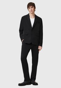 Black suit jacket with notch lapels and two front pockets, paired with matching trousers. Worn over a white crew neck shirt.