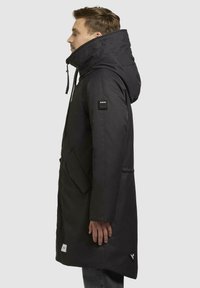 Sort parka med høj krave og justerbar hætte, med store sidelommer, et mærke på ærmet og en slank, lige silhuet.
