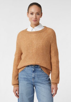 Femme portant un pull en tricot couleur camel sur une chemise blanche à col, associé à un jean bleu taille haute, se tenant devant un fond uni.
